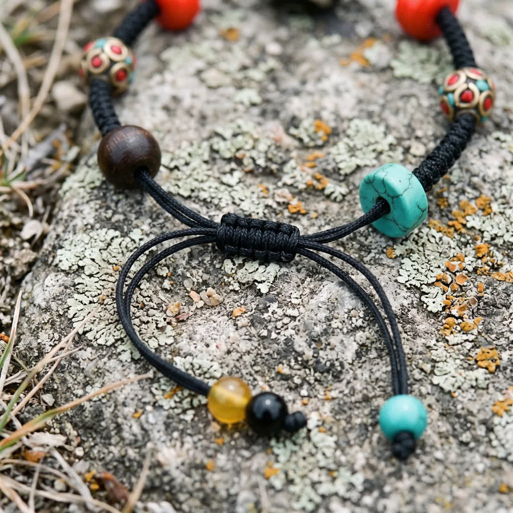 Close-up of the braided sliding knot where the cord tails meet, ending in yellow, black and blue-green beads, beside an agarwood bead and a blue-green disc