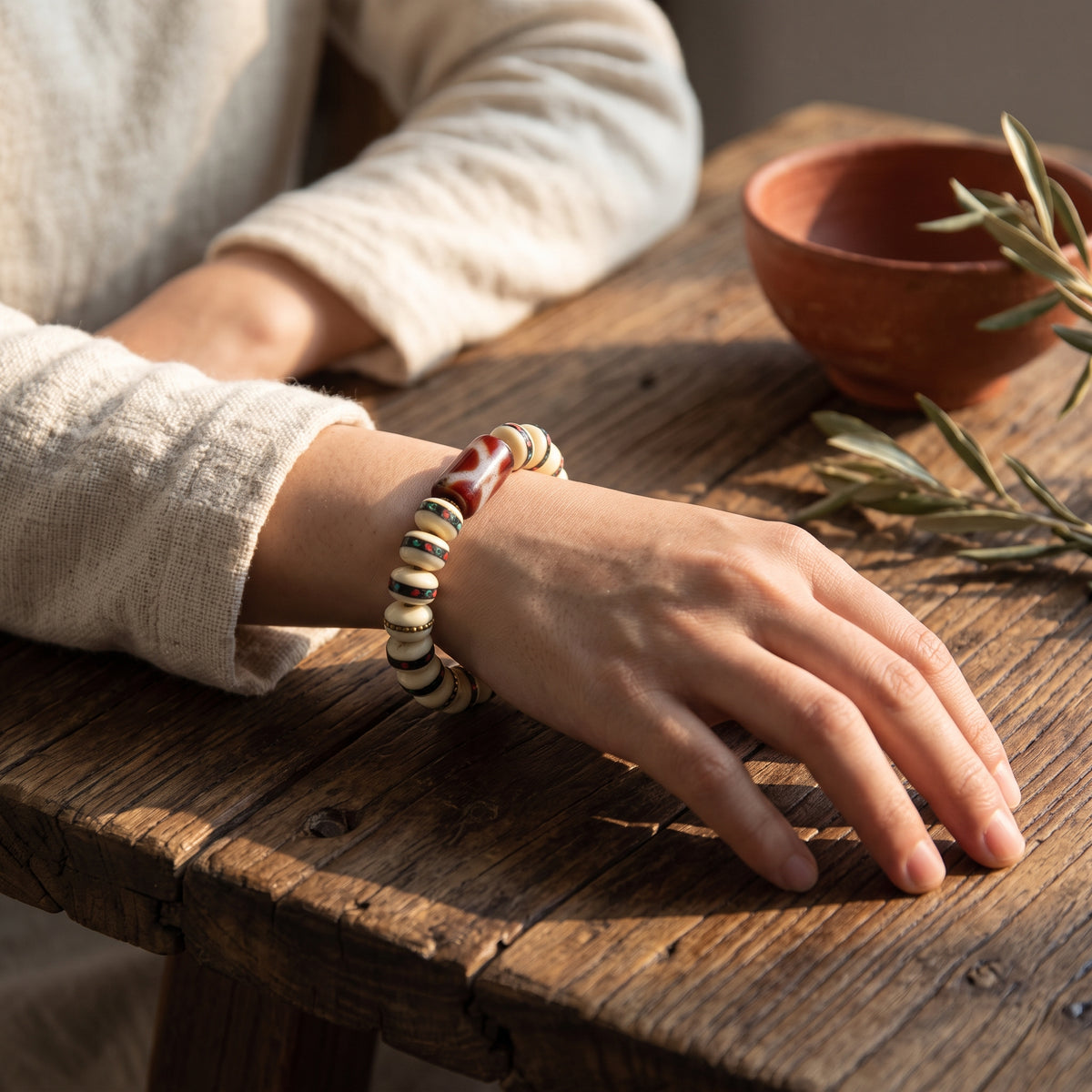 Wrist in a linen sleeve wearing a bone bead bracelet with a red-brown dzi bead, a clay bowl and olive leaves nearby