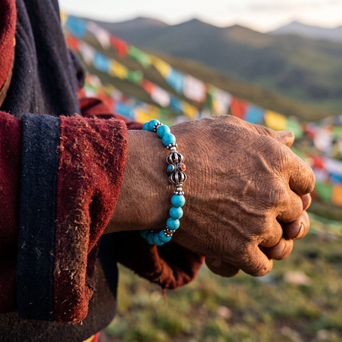 Weathered hand in a maroon sleeve wearing a turquoise and silver vajra bracelet, prayer flags on the hillside