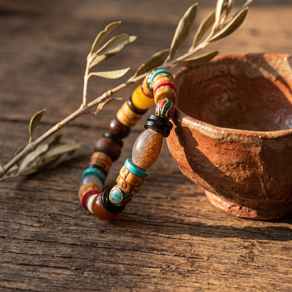 Multi-bead bracelet leaning against a rough clay bowl, an olive sprig and low morning light on weathered wood