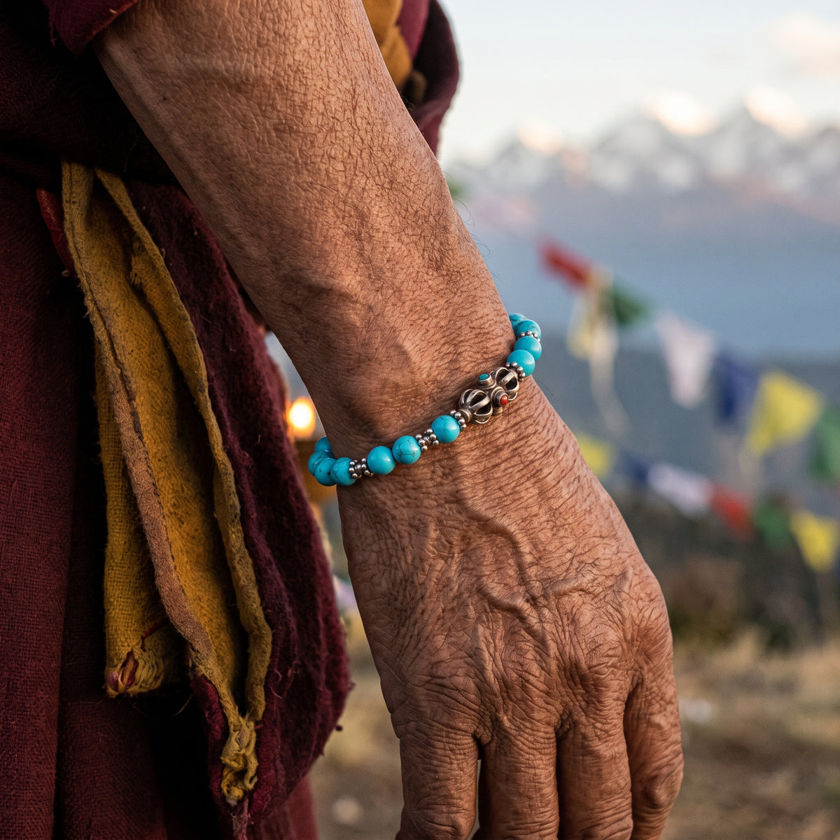 Wrist wearing a slim turquoise bracelet with a silver vajra, prayer flags and snowy peaks behind