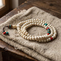 108-bead white yak bone mala coiled on undyed wool cloth, with red and turquoise accent beads and an inlaid brass guru tube
