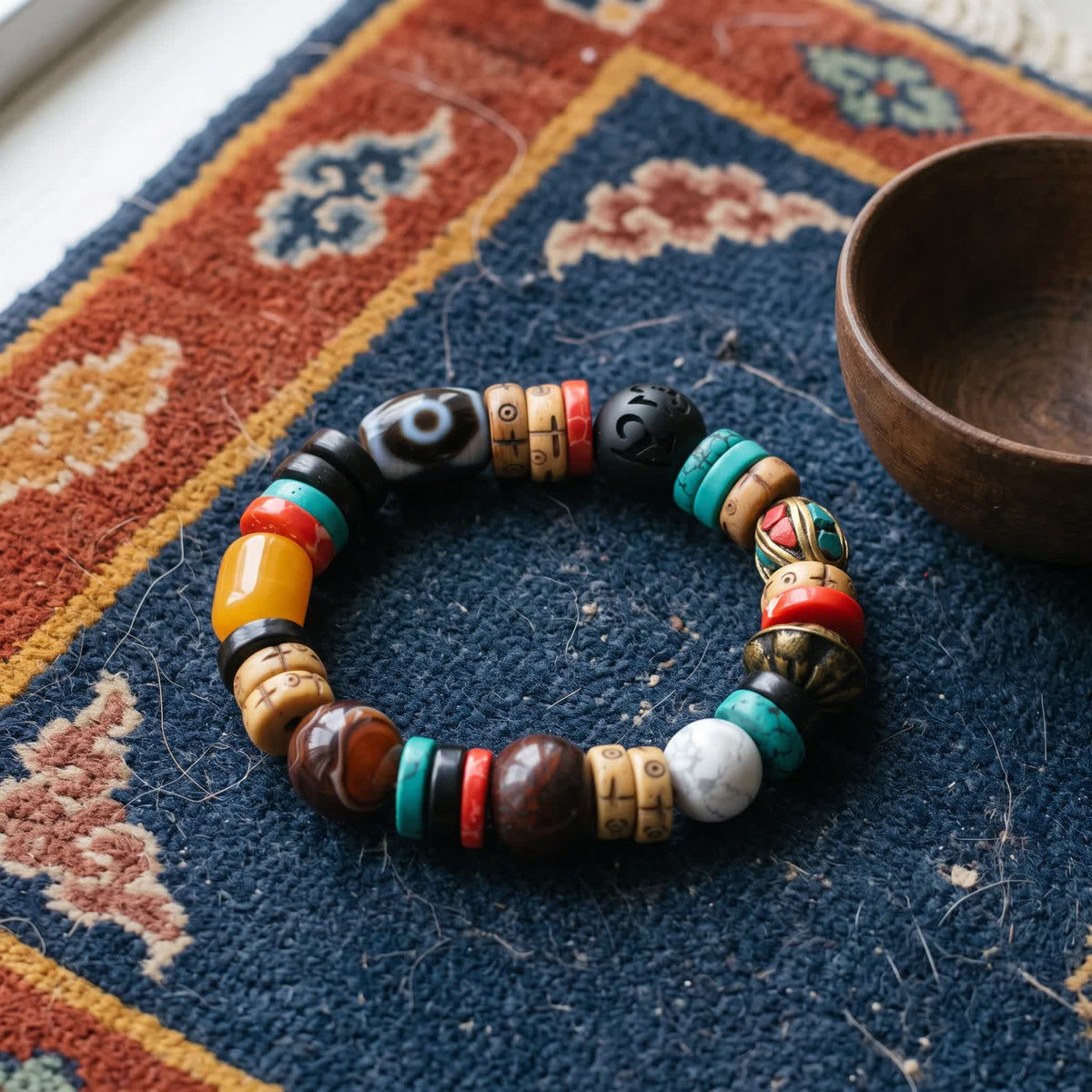 Dzi bracelet with carved bone, turquoise, red and brass beads on an indigo Tibetan khaden rug beside a wooden bowl