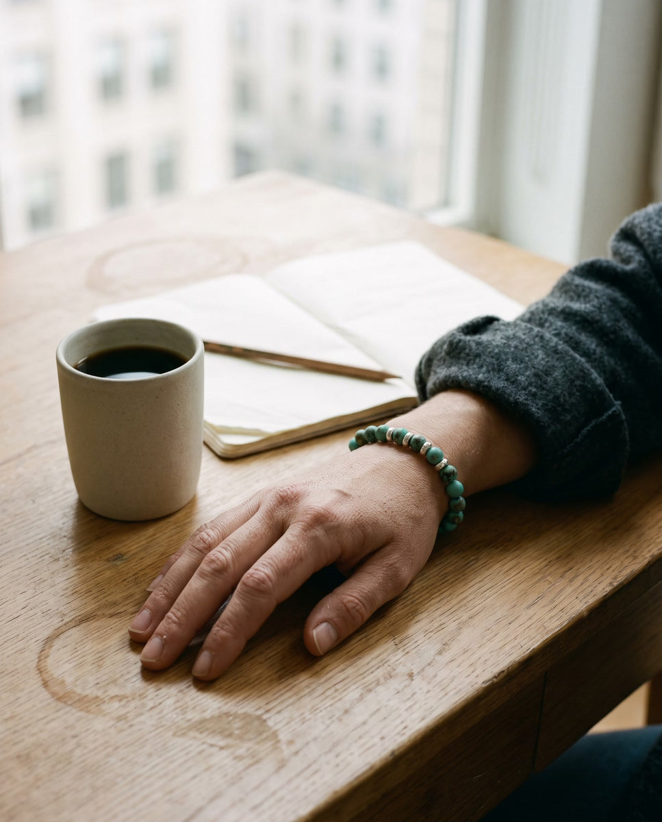 A hand wearing a turquoise and silver bead bracelet on a wooden desk beside black coffee and an open notebook