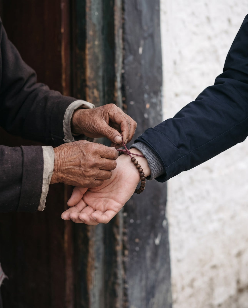 Old weathered hands tying a strand of dark wooden beads onto a younger wrist in a navy jacket sleeve, in a Lhasa doorway