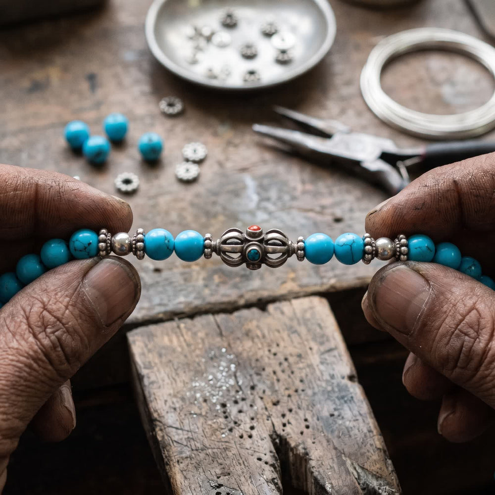 Weathered hands holding the bracelet's silver vajra between small silver spacers and blue turquoise beads at a wooden workbench